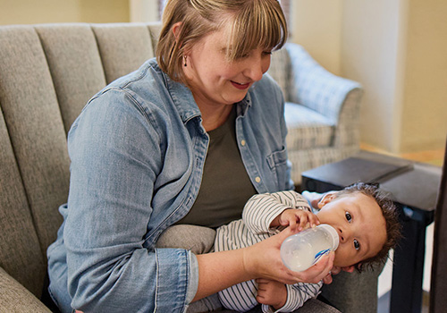 A mother wearing a blue denim shirt looks down at her young baby in her arms. She is holding her son on his side and feeding him a bottle of formula as part of therapy.
