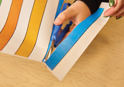 A hand using blue adaptive loop scissors cuts through paper with color guidelines as a part of therapy.