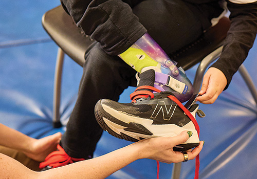 A child is seated in a chair wearing a brightly colored Ankle-Foot Orthosis (AFO). With the assistance of his female therapist, he is using a shoe horn to put his black sneaker on his foot.
