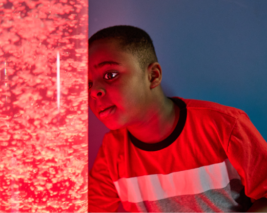 A boy is looking very closely at a sensory bubble tube that is giving off a red glow to everything around.