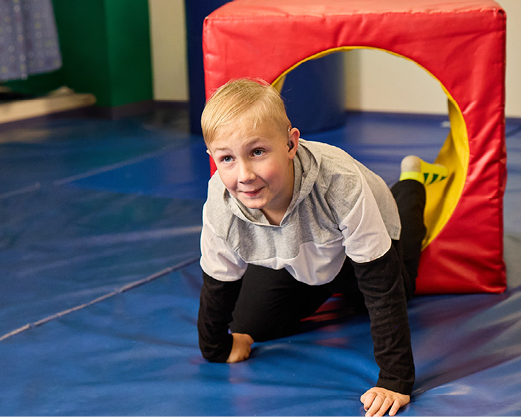 A 10-year-old boy with blonde hair wearing a hearing aid is crawling through a red play tunnel in an occupational therapy gym.