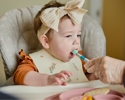 A baby girl wearing a large tan bow on her head is chewing on a Nuk brush as part of occupational therapy.