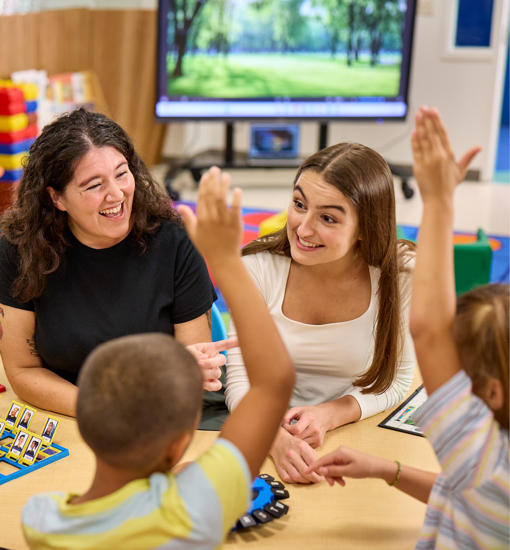 two teachers smiling while engaging with young children with their hands raised in a colorful classroom.