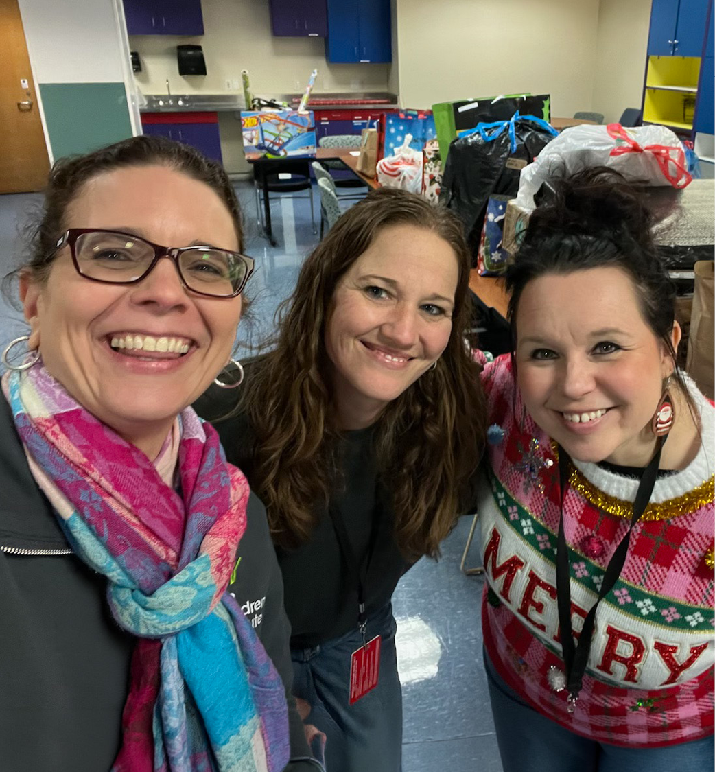 Three woman wearing colorful holiday sweaters and scarves are standing at the front of a classroom full of donated gifts.
