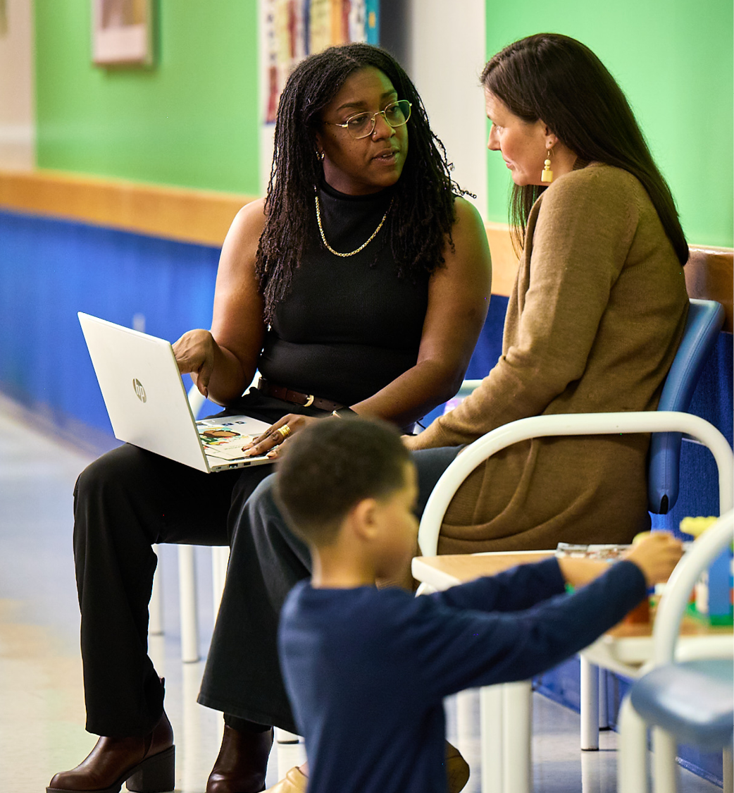 A female therapist is sitting in a hallway next to a mother discussing information shown on a silver laptop. a young boy wearing a blue shirts plays in the foreground.