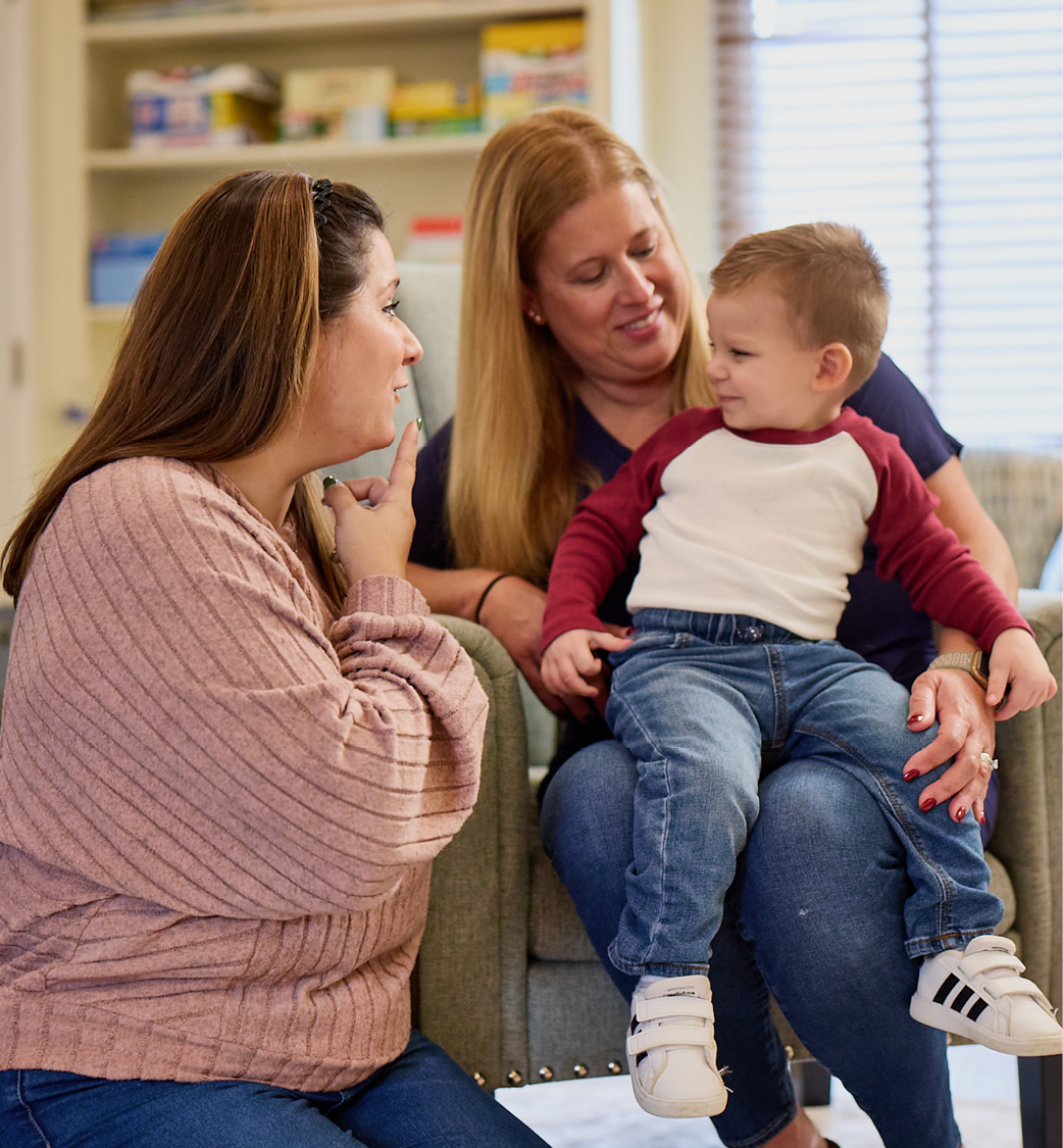 A young boy wearing a white shirt with long red sleeves sits on his mom's lap as a female therapist wearing a pink top is kneeling at their side. The therapist is pointing to her mouth showing this is an at home speech session.
