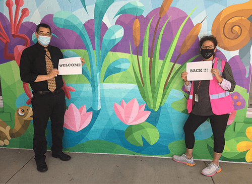 Image of two team members wearing safety masks, socially distanced holding "Welcome Back" signs