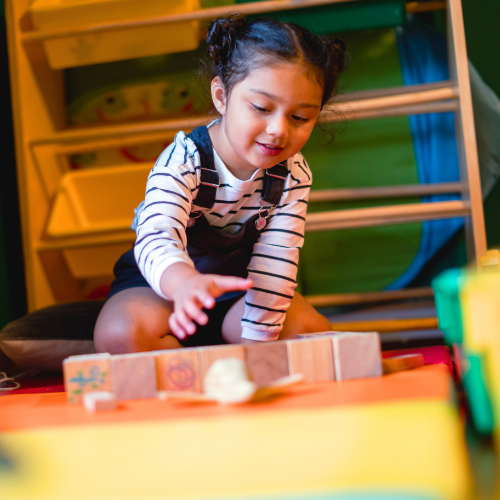 A young girl playing wooden blocks on a colorful rug in a classroom