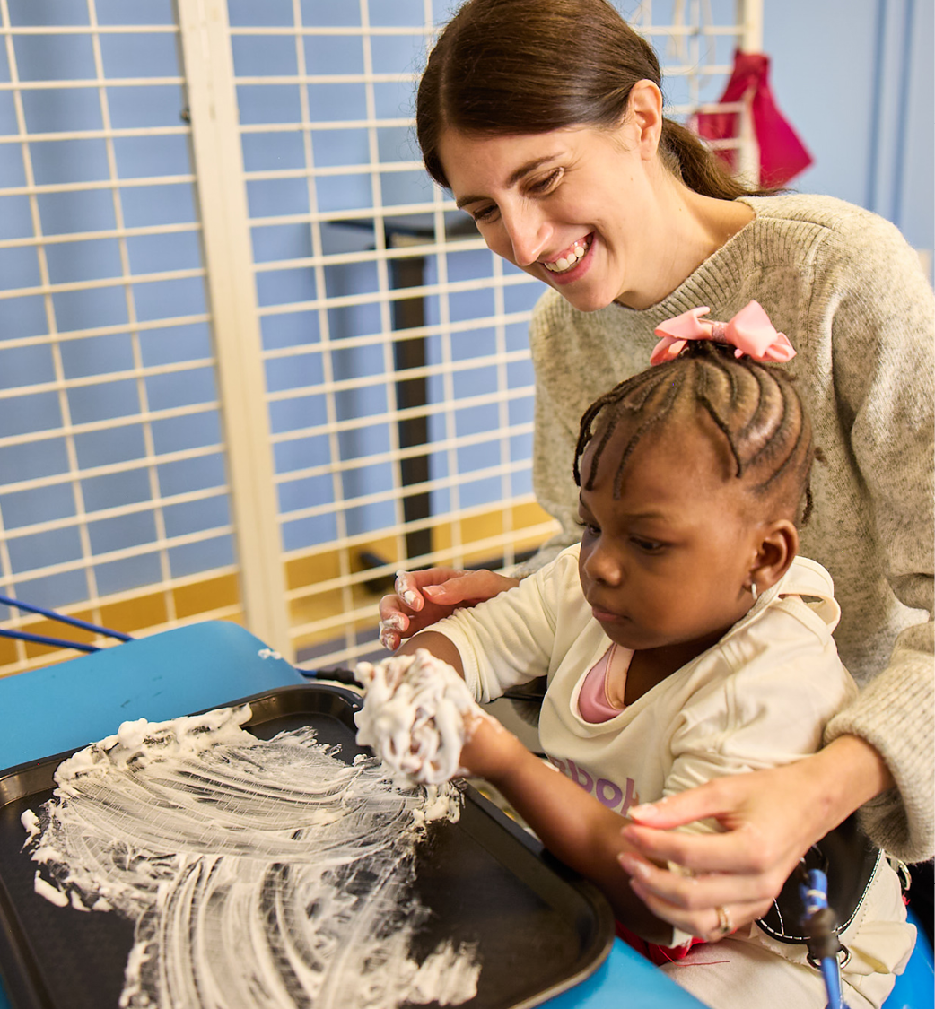 A female therapist sits closely behind a young girl with black braids and a pink bow in her hair. The girl is having a therapy session where she is playing in a tray of white shaving cream, working on sensory input as she works her muscles in a device
