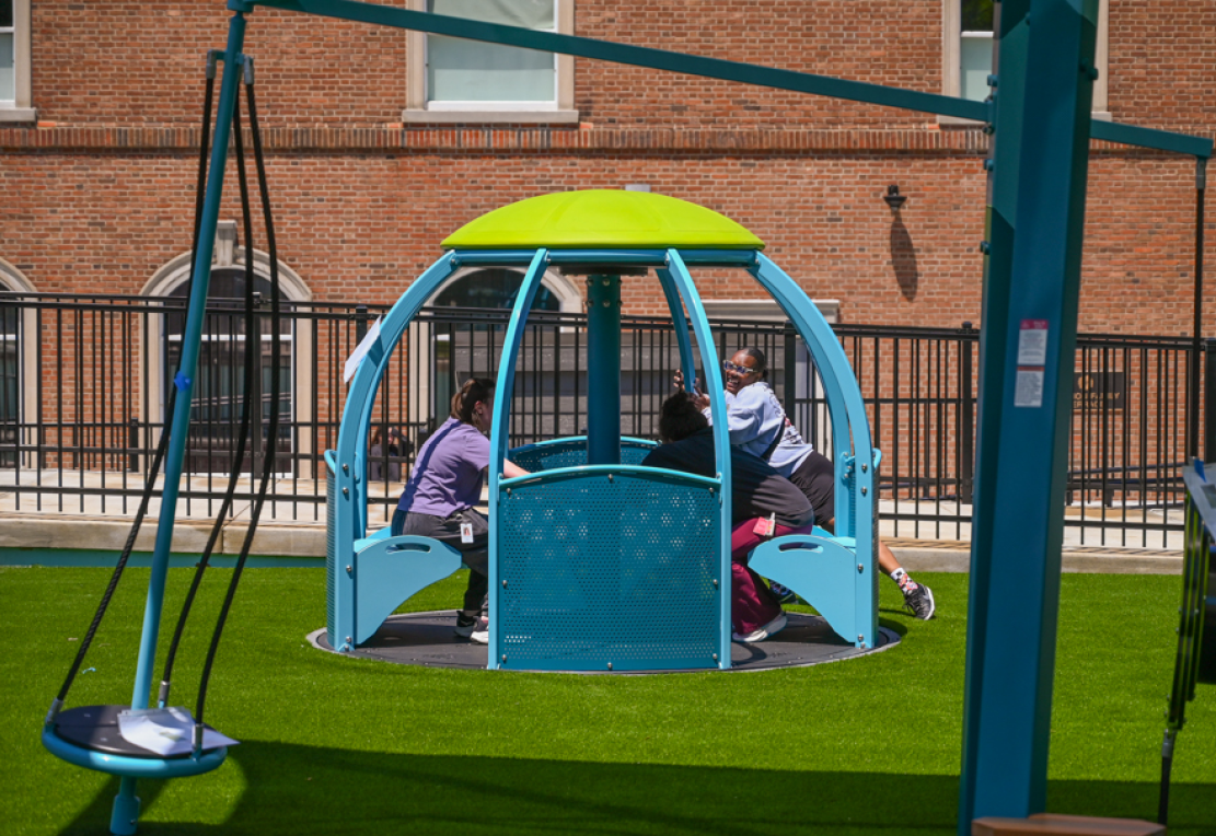 Three teachers try out an accessible blue and green merry-go-round on a new playground. There is a red brick building in the background.