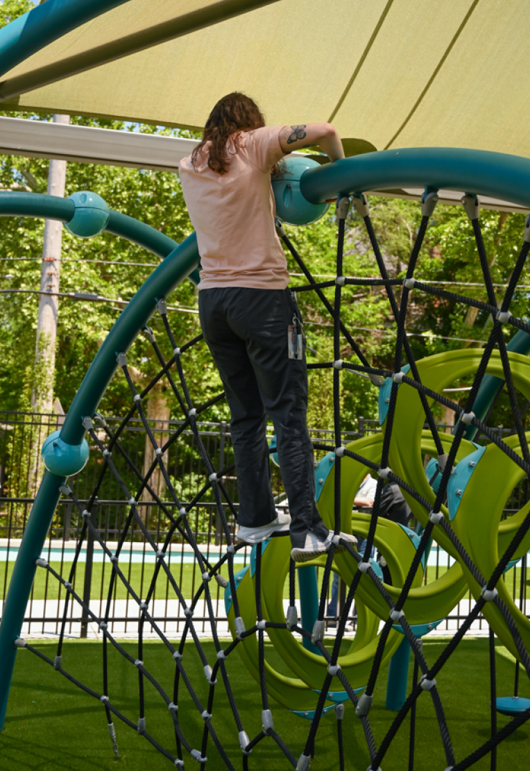 A teacher climbs across a black cargo net on a colorful blue and green jungle gym. There is a light green canopy above her and green trees in the background.