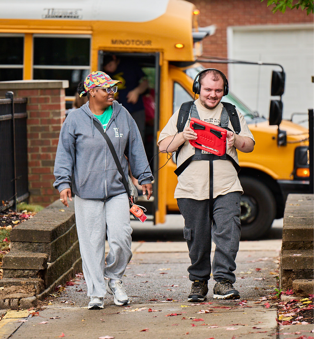 A female teacher wearing a colorful ballcap and gray zip up sweatshirt is walking with an older student who is holding a red tablet in his hands for communication. The pair are walking in from a school bus to start the school day.